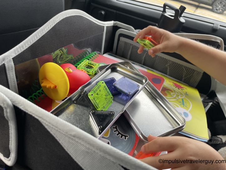 A child's hands playing with translucent colorful magnetic tiles on a metal lap desk tray inside a car, with a toy organizer nearby.