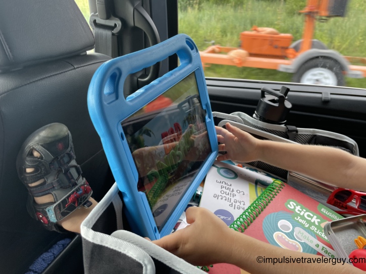 View from the back seat of a car showing a child watching an iPad in a blue protective case while resting their hands near a reusable sticker pad.