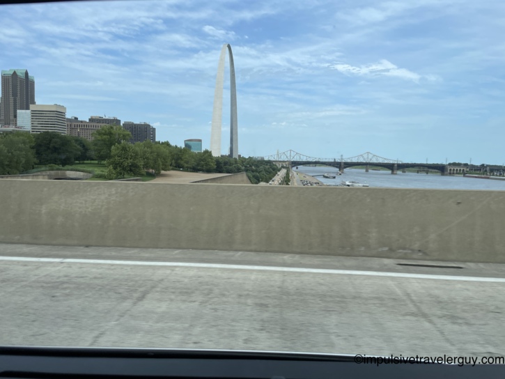 View from a moving vehicle driving across a highway bridge, showing the Gateway Arch and the downtown St. Louis skyline in the distance.