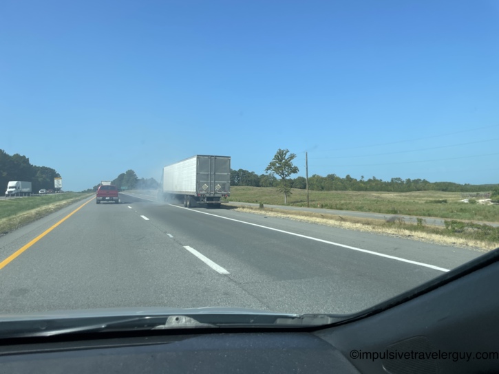 View from a moving vehicle on a highway showing a white semi-truck smoking from tire issues and pulling onto the grassy shoulder due to the heat.