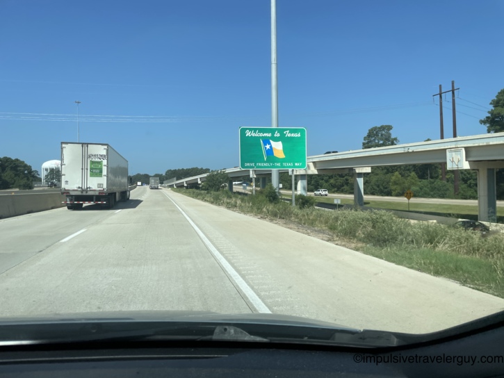 Driving on a highway approaching a green 'Welcome to Texas' sign featuring the state flag posted on the shoulder.