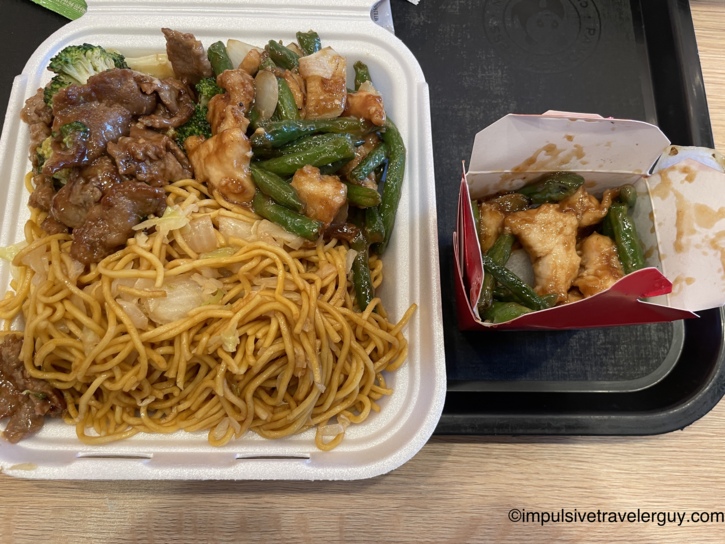 A Panda Express takeout meal showing a styrofoam container filled with chow mein, broccoli beef, and orange chicken, alongside a smaller cardboard box of string bean chicken.