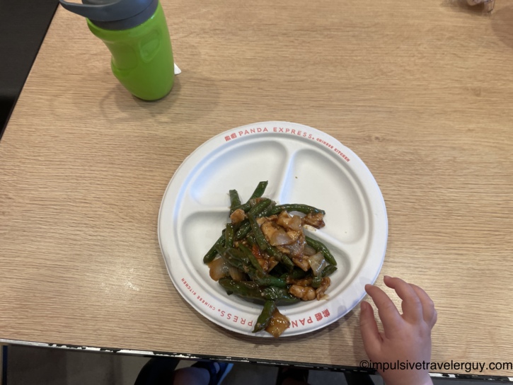 A child's hand reaching toward a portion of string bean chicken served on a divided Panda Express paper plate, resting next to a green sippy cup.