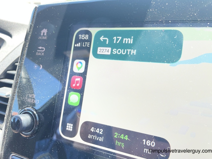 Close-up of a car dashboard navigation display showing a suggested Google Maps route requiring a left turn onto a rural Farm-to-Market road.