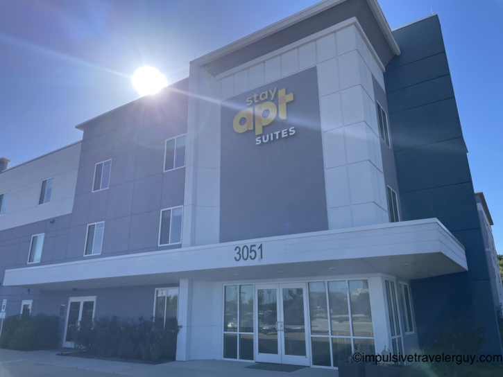 Exterior of a modern stayAPT Suites hotel building in Bryan featuring a grey and dark blue facade under a bright sun.