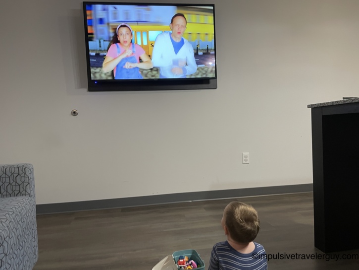 A young child sitting on the floor looking up at a wall-mounted flat-screen TV playing a Ms. Rachel video to decompress.
