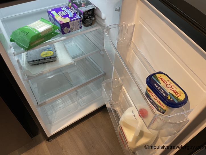 An open full-size hotel fridge stocked with essential snacks and drinks including string cheese, Uncrustables, yogurt, and milk.