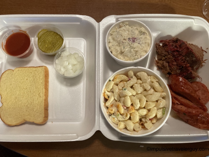 A C&J BBQ meal in a styrofoam container, featuring sliced smoked sausage, chopped brisket, macaroni salad, potato salad, and a slice of plain white bread.