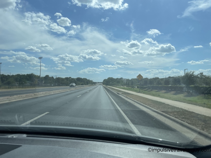 View from inside a moving vehicle driving down a highway under a bright blue sky filled with scattered clouds.