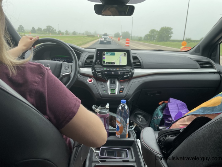 View from the back seat of a car between two child car seats, showing Victoria driving with the dashboard navigation screen visible.