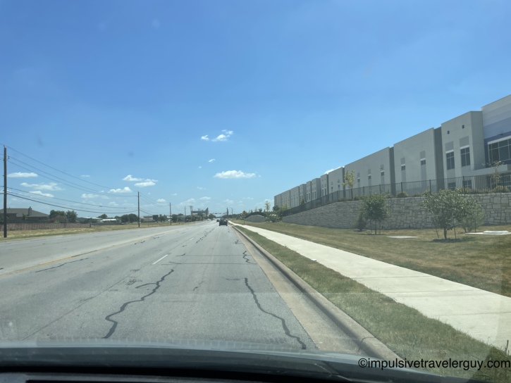 View from a moving vehicle driving down Sunrise road, passing a large, newly built complex elevated by a stone retaining wall.