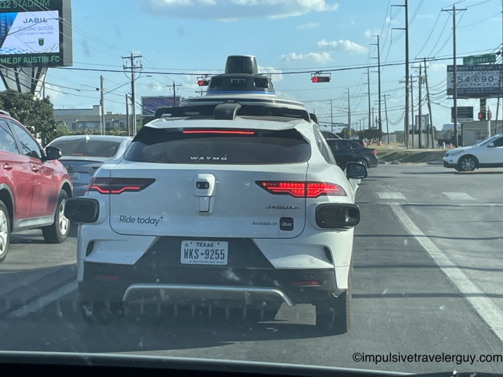 View from a vehicle stopped behind a white Waymo autonomous driverless vehicle equipped with a large roof-mounted sensor dome.