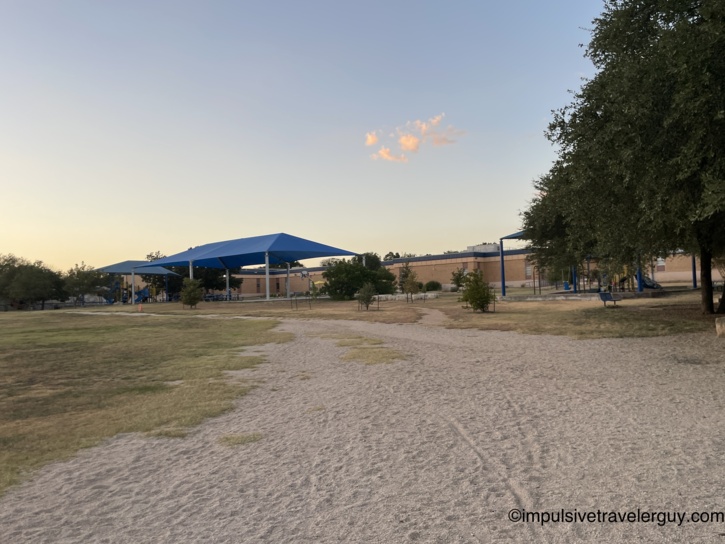 A park playground area at dusk featuring blue shade canopies covering the play equipment to keep it cooler in the Texas heat.