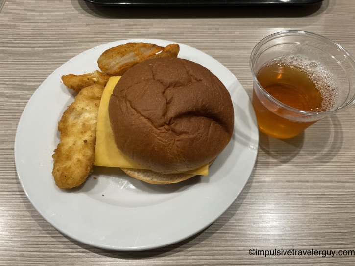A hotel breakfast plate holding a simple sandwich, chicken tenders, and potato wedges next to a cup of juice.