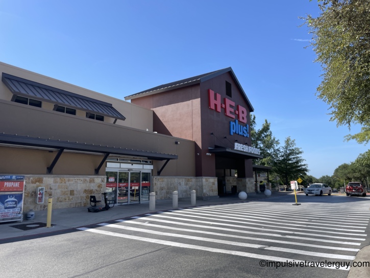 Exterior daytime entrance of an H-E-B plus! grocery store featuring a brown facade and red and blue store signage.