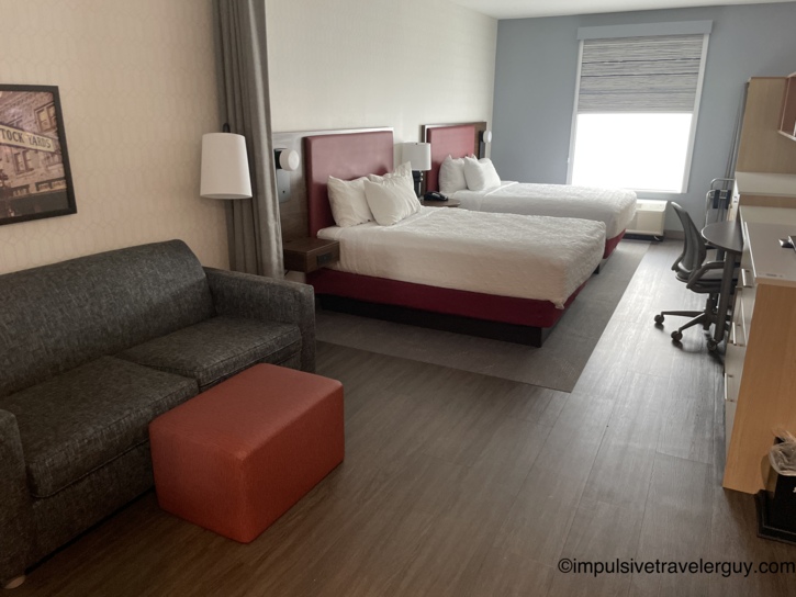Interior of a Fort Worth Home2Suites hotel room featuring two beds with red headboards, a grey sofa, and a red ottoman.