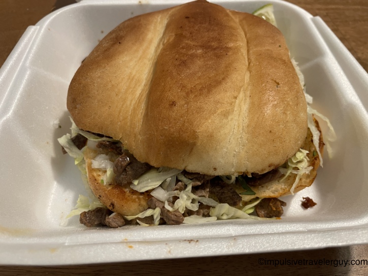 A Mexican torta sandwich filled with chopped meat and shredded lettuce, resting inside a white styrofoam takeout container.