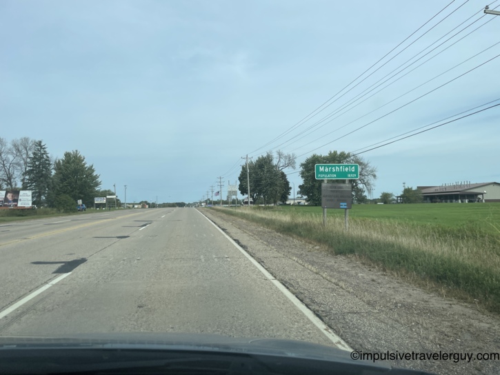 A view through a car windshield driving down a two-lane highway, approaching a green city limit sign on the right side of the road that reads 