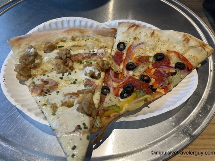 Two large slices of pizza on a white paper plate resting on a metal pizza pan. One slice features a white sauce base topped with chicken, bacon, and chives. The other slice is topped with tomato sauce, pepperoni, sliced bell peppers, red onions, and black olives.