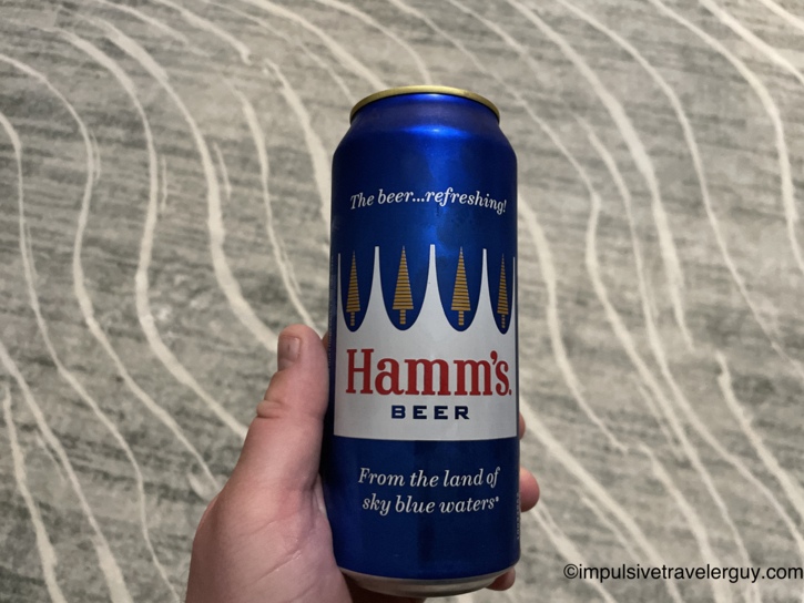 A person's hand holding a cold, blue can of Hamm's Beer against a patterned background.