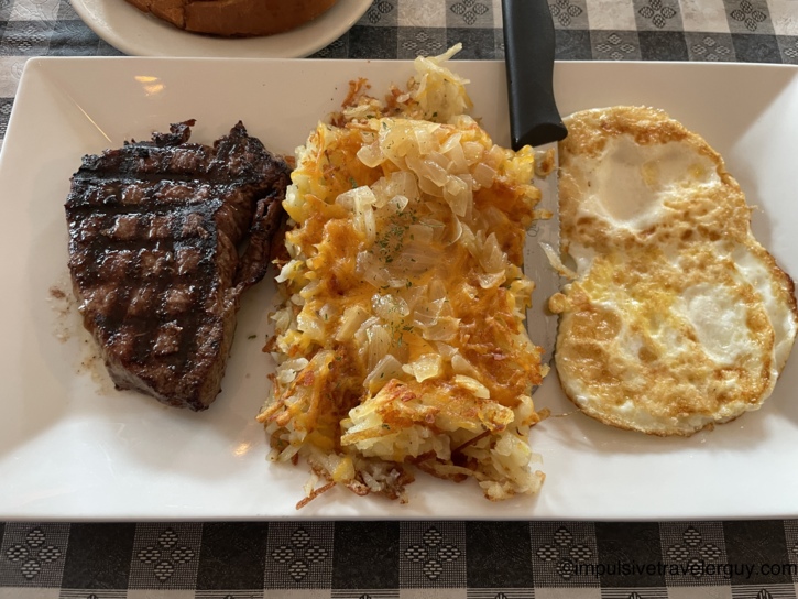 A rectangular white plate featuring a grilled steak, a large scoop of hashbrowns topped with melted cheese and diced onions, and two fried eggs.