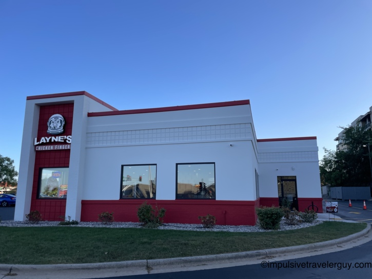 The exterior of a Layne's Chicken Fingers restaurant on a sunny day, featuring white walls, a red brick base, and the restaurant's logo above the front entrance.