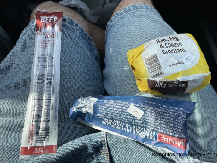 A view looking down at a person's lap in a car, showing a packaged Wenzel's Farm beef stick, a packaged Minh egg roll, and a yellow-wrapped ham, egg, and cheese croissant.